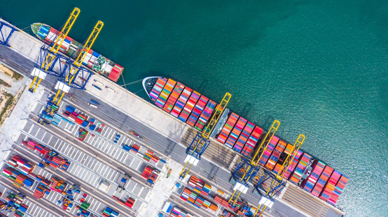 Overhead perspective of a container ship at a port, showcasing the ship's cargo and the busy dock area.
